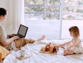 mother using laptop while cute little girl playing on bed at home
